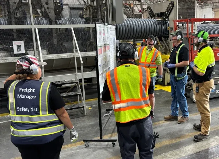 A trainer briefs workers in hard hats and safety vests on a plant floor