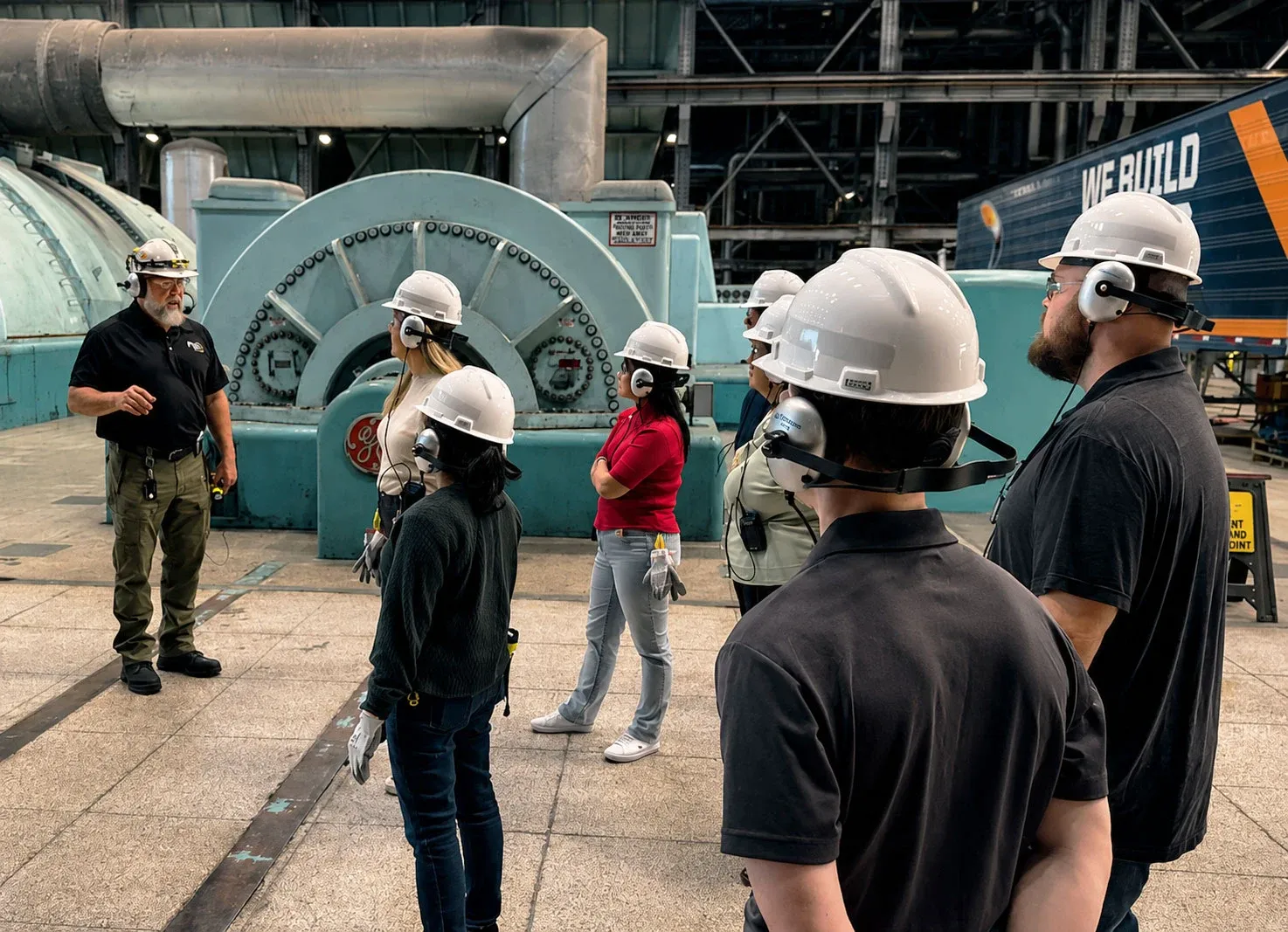 A tour group in hard hats and headsets listens to a guide beside a turbine