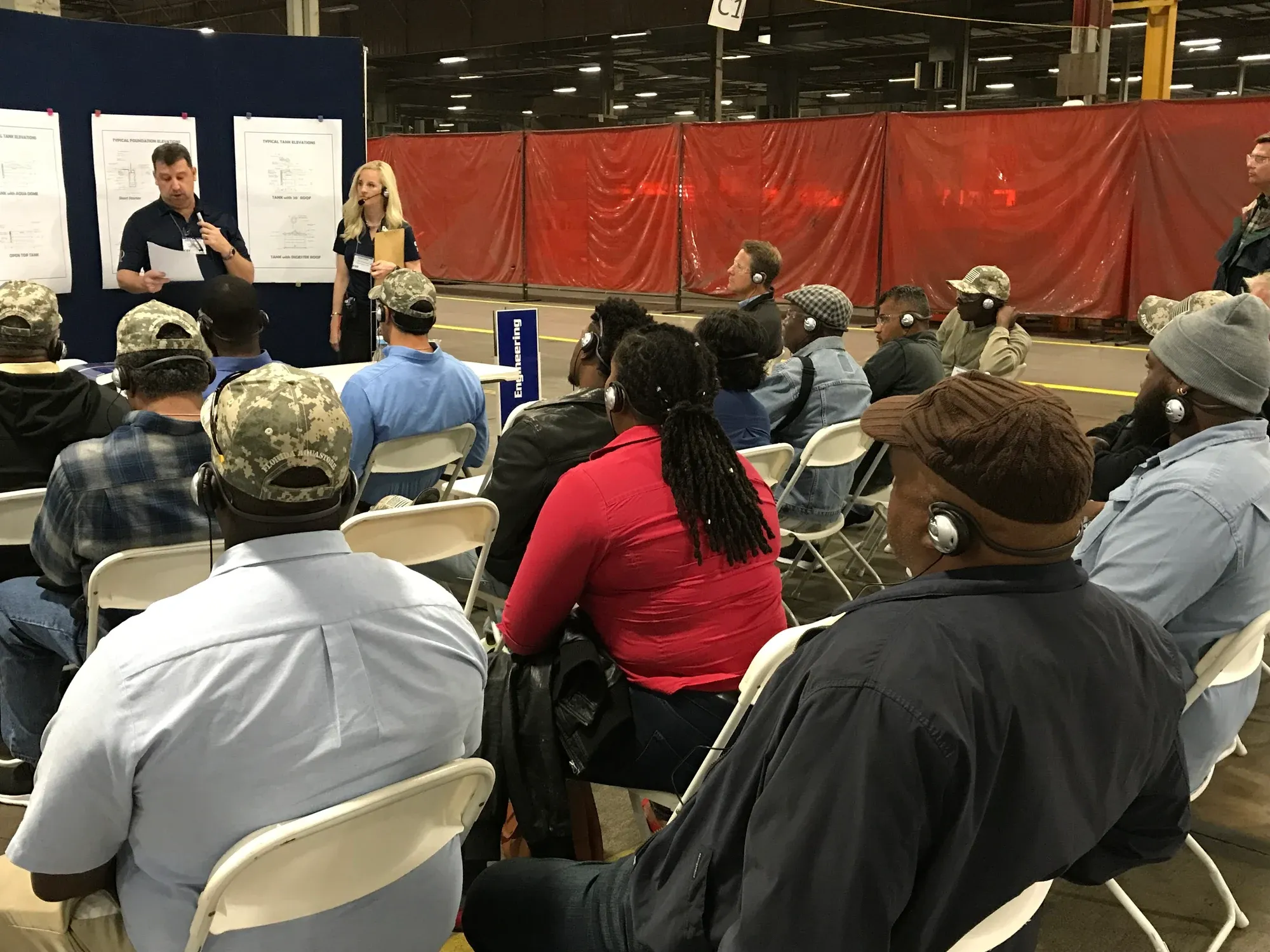 Seated audience wearing headsets, listening to presenters in a plant
