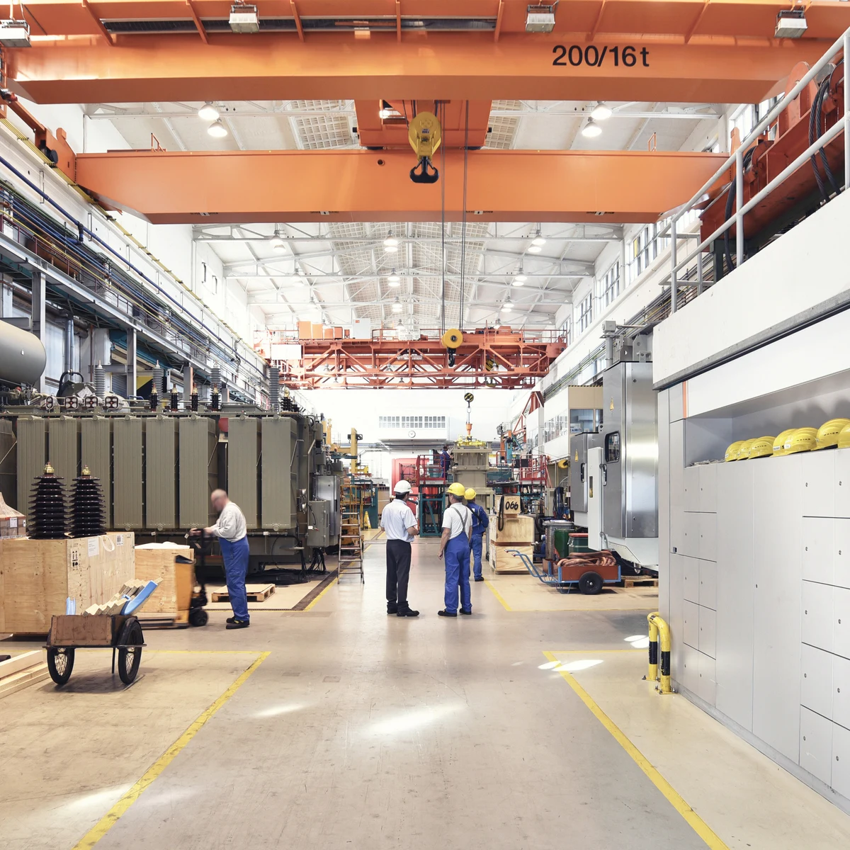 Large industrial factory hall with an overhead crane, machinery and workers along a lit aisle