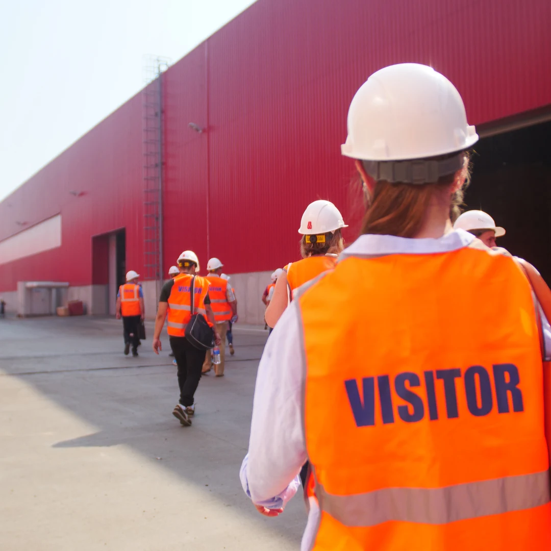 Person in a white hard hat and orange vest marked Visitor walking with a group outside an industrial building