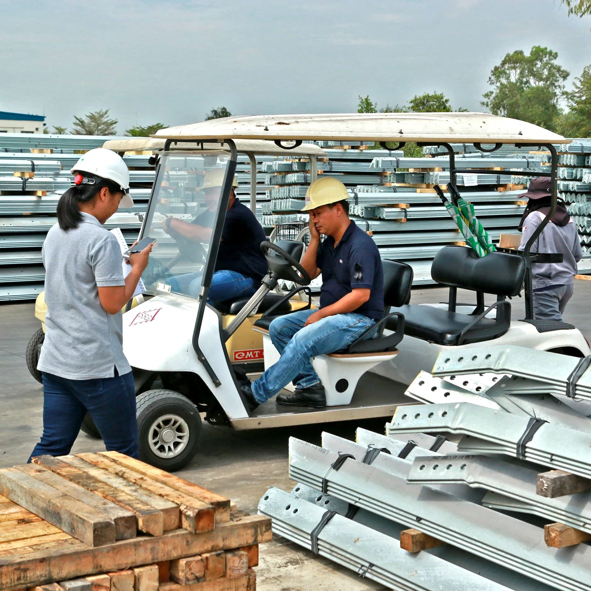 Workers in hard hats talking beside a utility cart in an outdoor yard stacked with steel materials