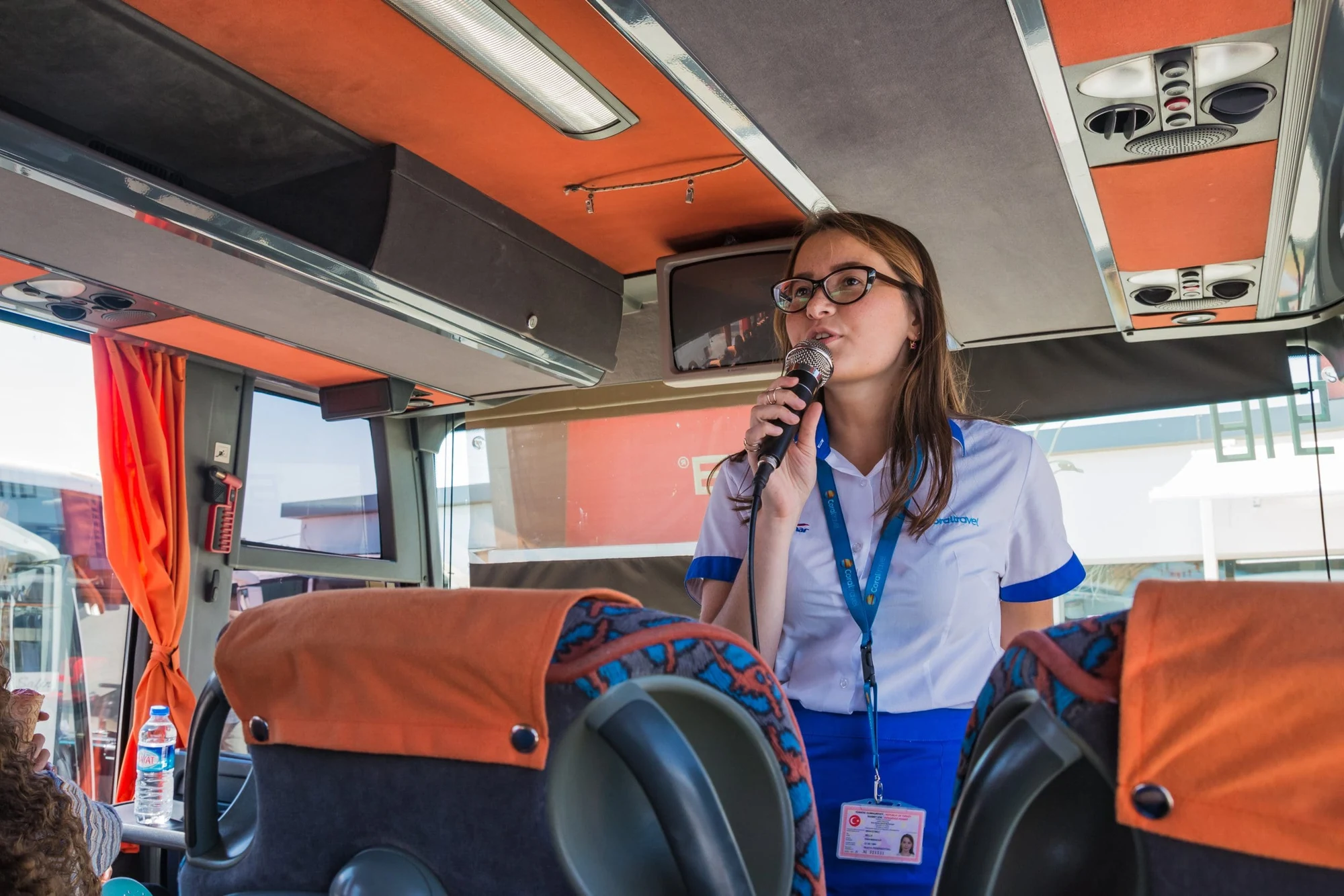 Tour guide with a lanyard speaking into a microphone at the front of a tour bus