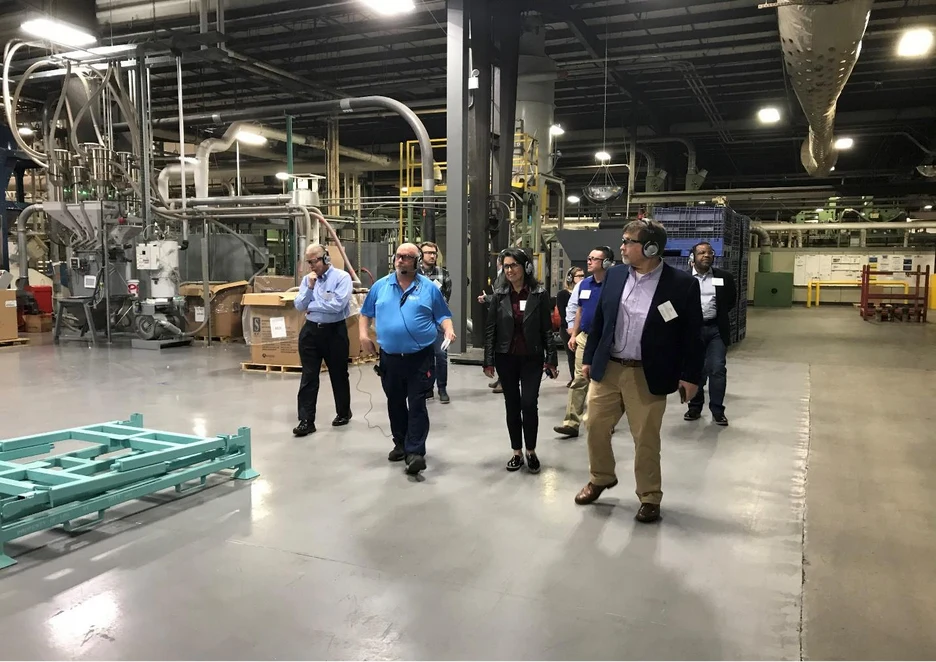 Tour group walking across a large manufacturing plant floor past machinery