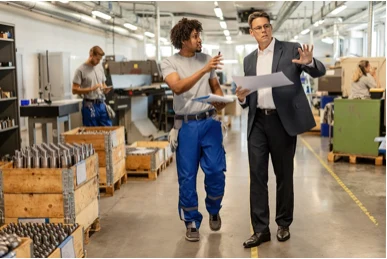 Two men talking and gesturing as they walk through a factory floor past machinery and parts crates