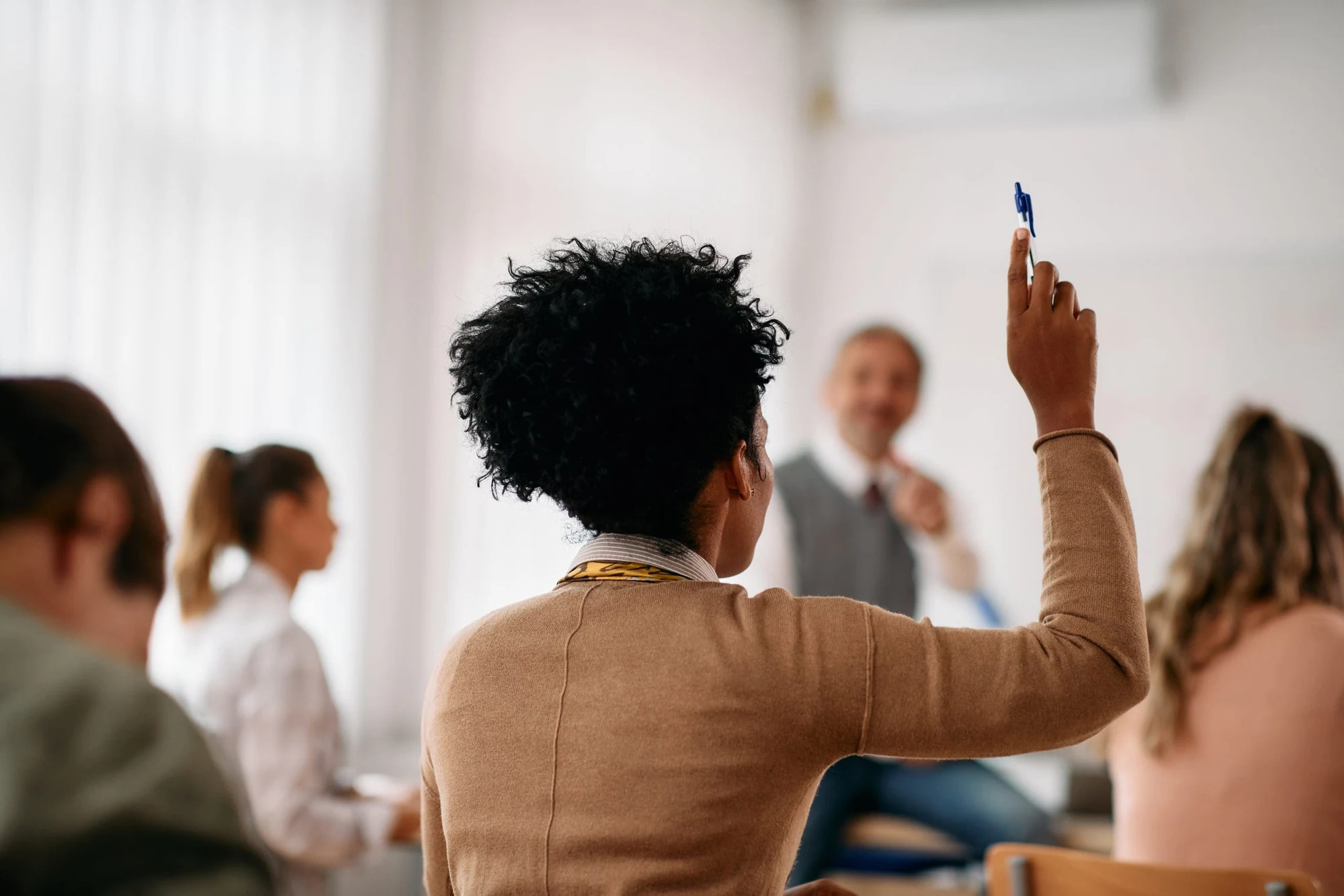 Woman in a classroom or seminar raising her hand holding a pen, with other attendees in the background