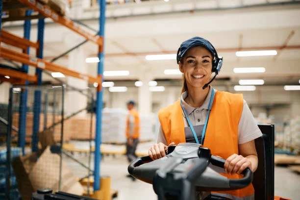 Smiling warehouse worker in an orange vest and headset driving a forklift