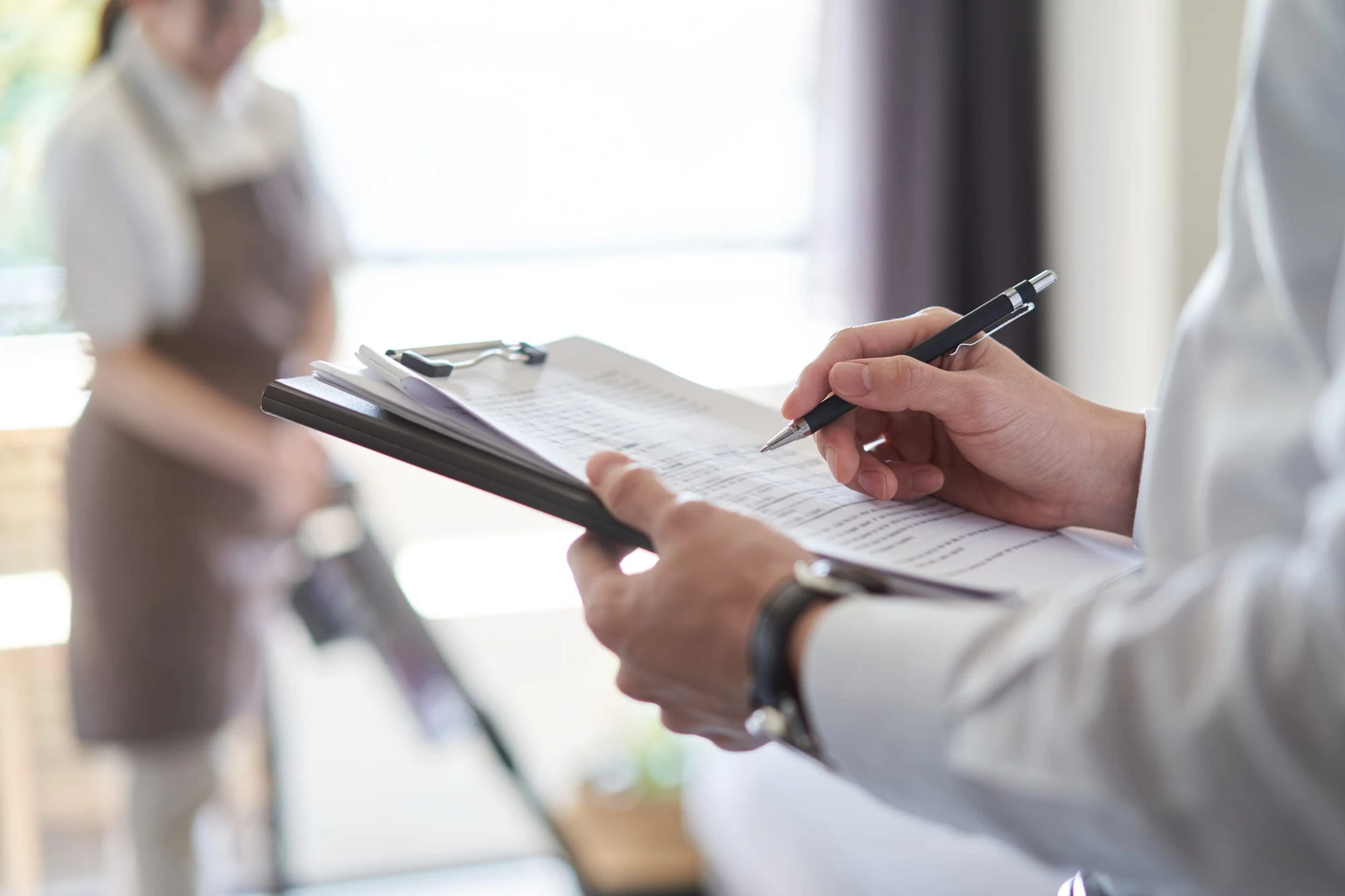 Person holding a clipboard and pen checking a list, with staff in the background