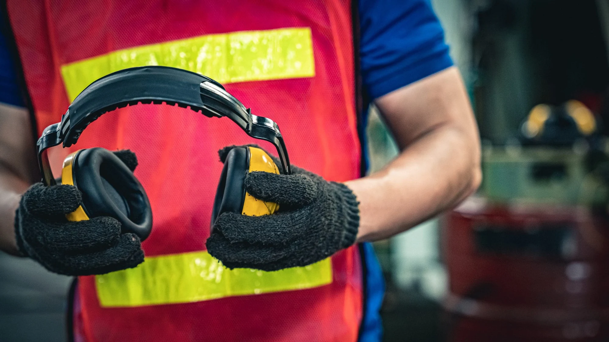 Worker in a high-visibility vest holding a pair of yellow and black ear muffs in gloved hands