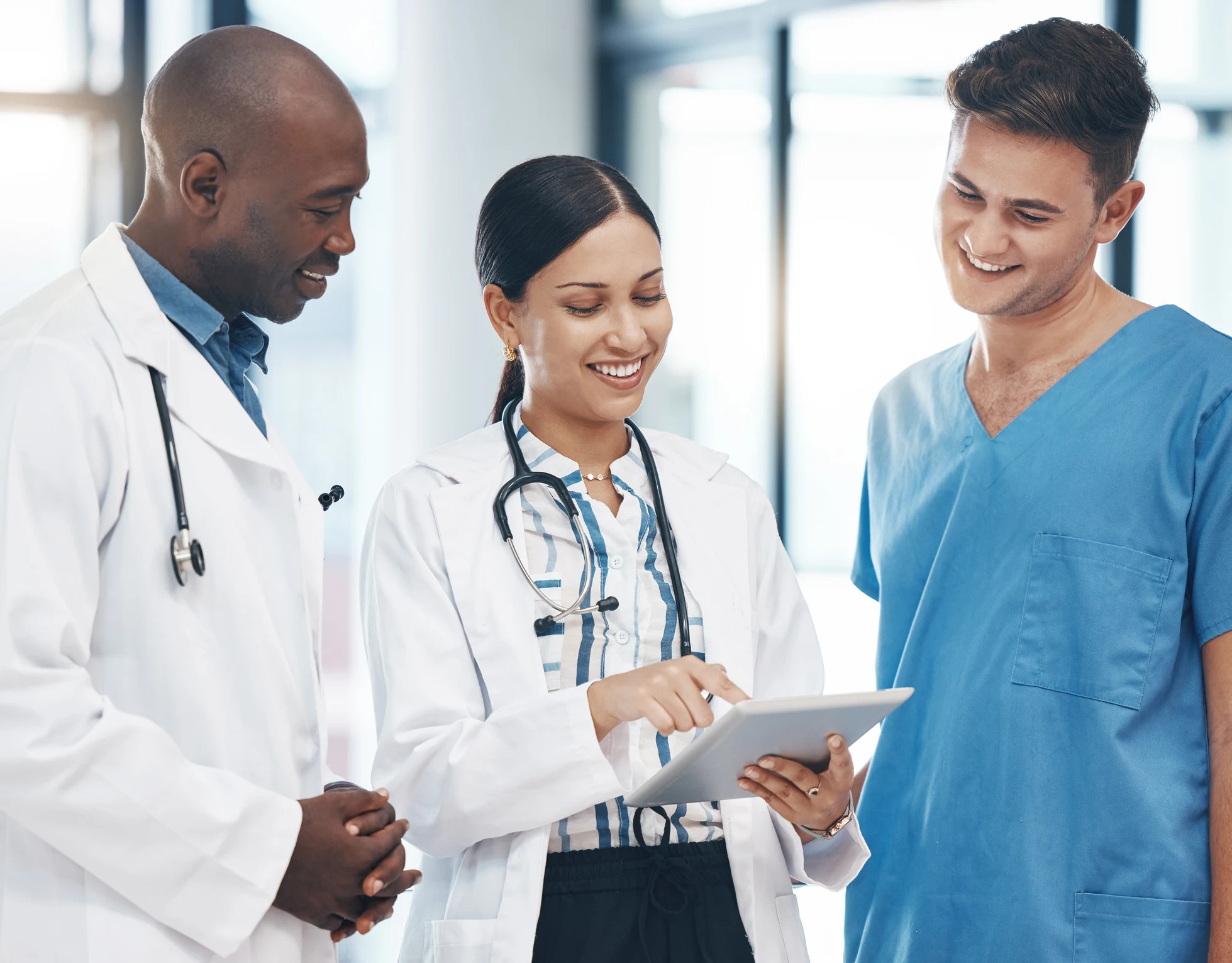 Smiling doctors and a nurse in scrubs looking at a tablet together in a hospital hallway