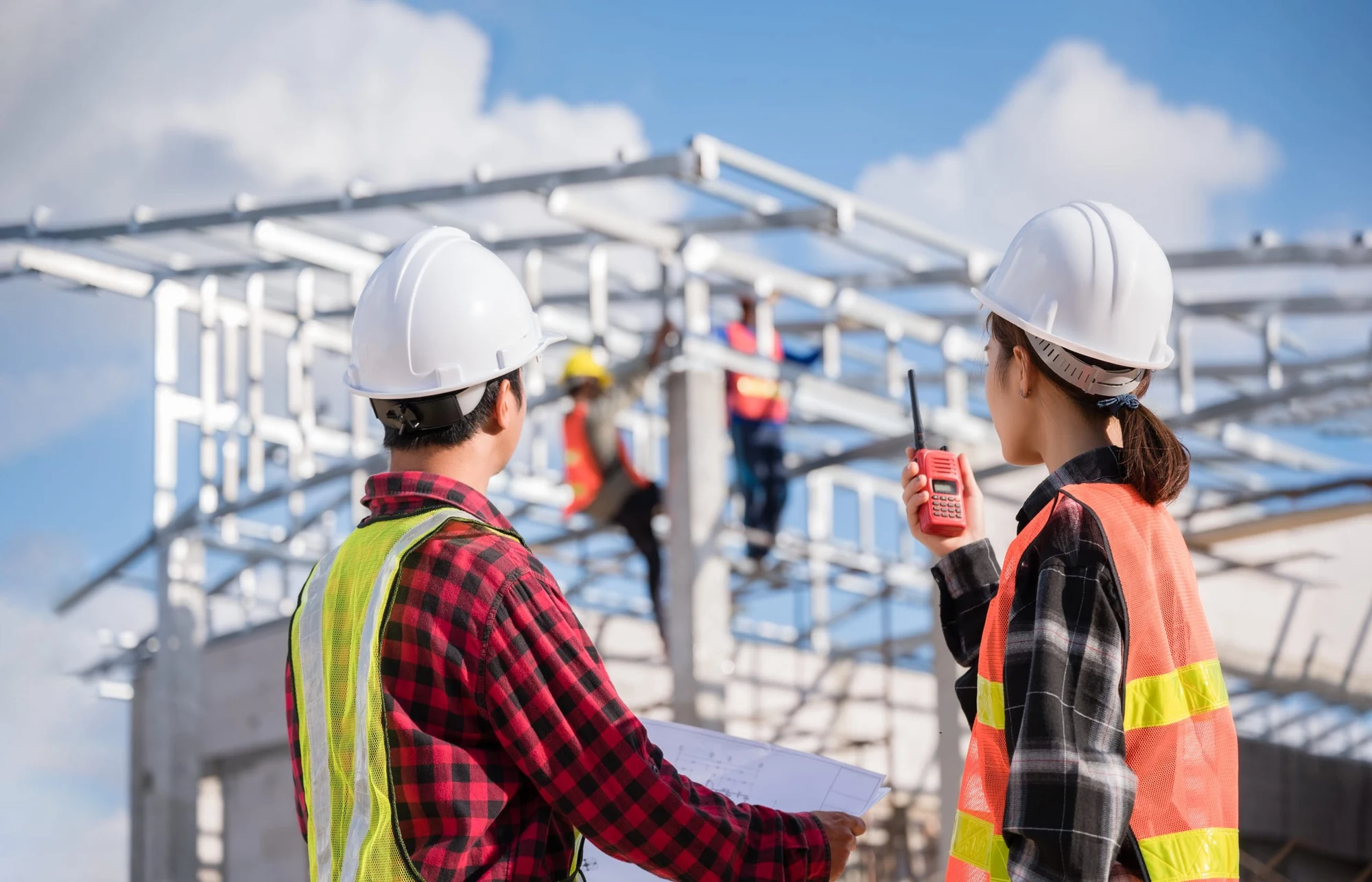 Two workers in hard hats and safety vests at a construction site, one holding a two-way radio