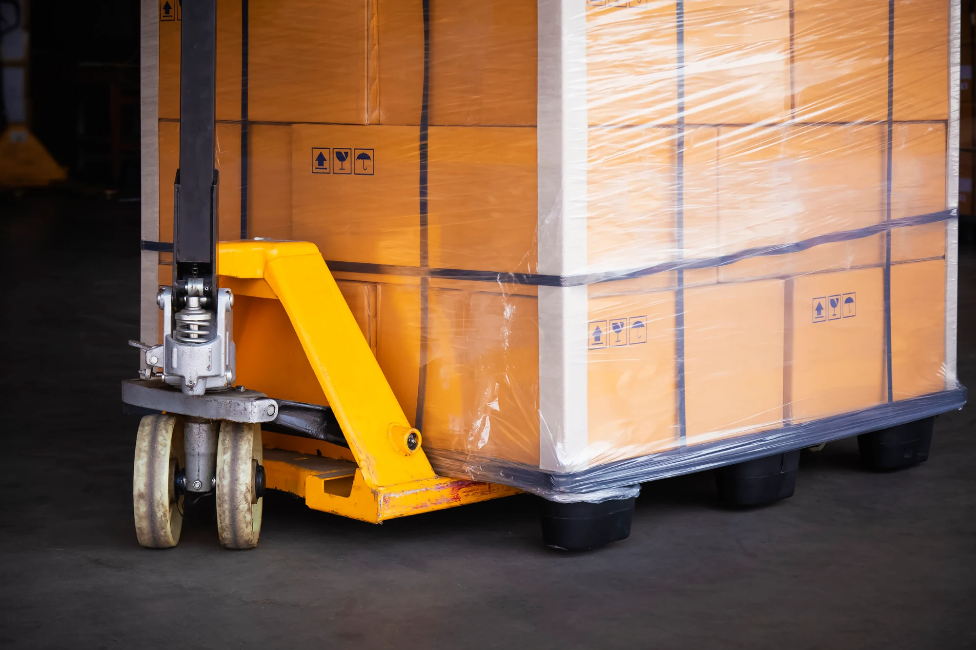 Yellow pallet jack under a pallet of shrink-wrapped cardboard boxes