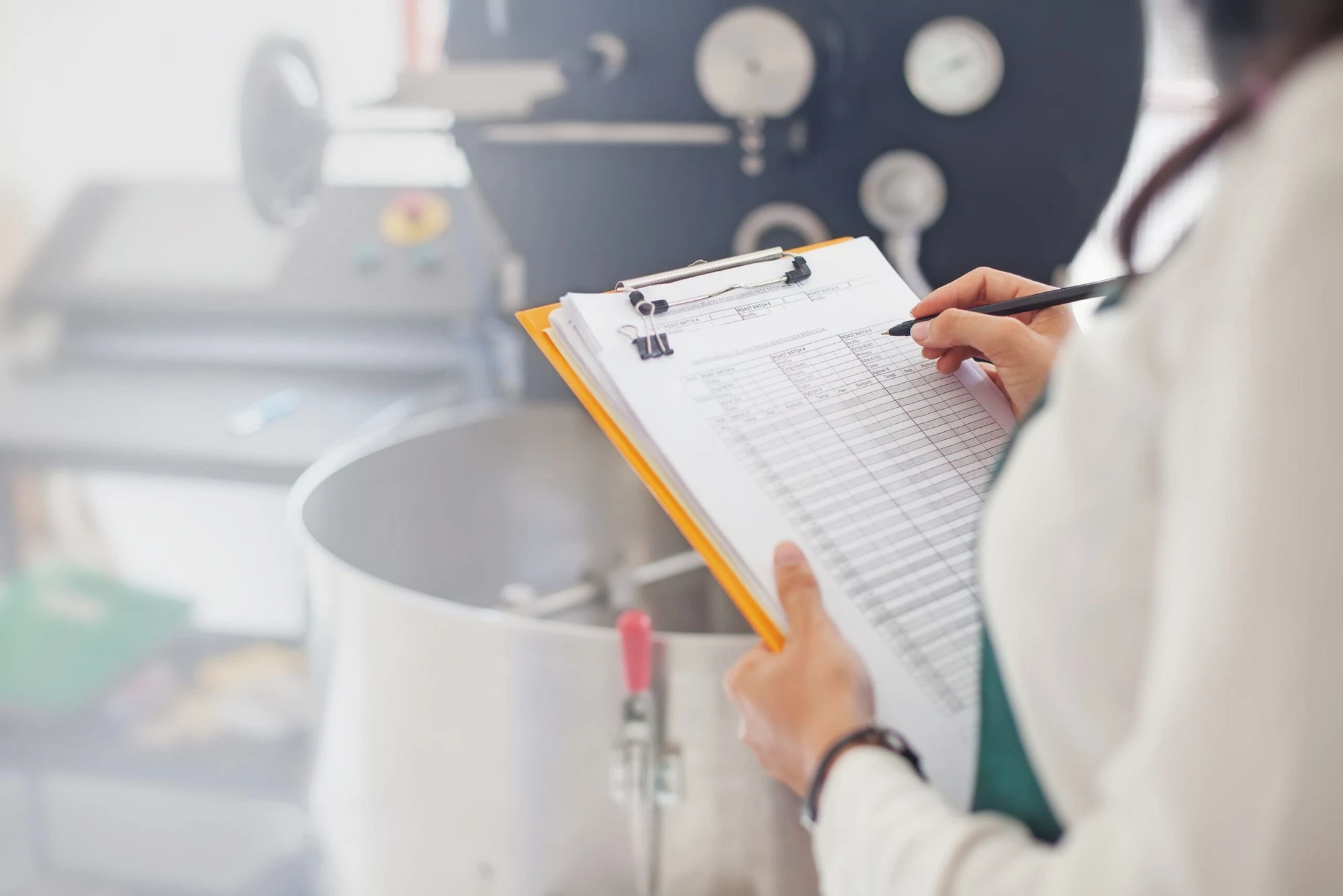 Person in a white coat writing on a clipboard checklist next to industrial equipment