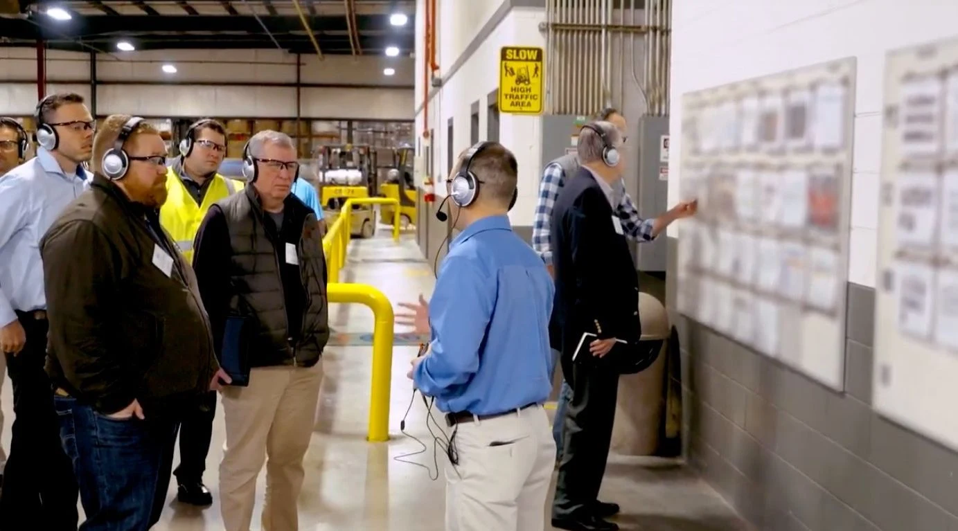 Tour group wearing headsets and safety glasses looking at displays inside a factory