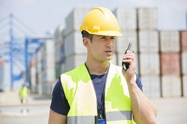 Worker in a yellow hard hat and safety vest speaking into a two-way radio at a shipping container yard