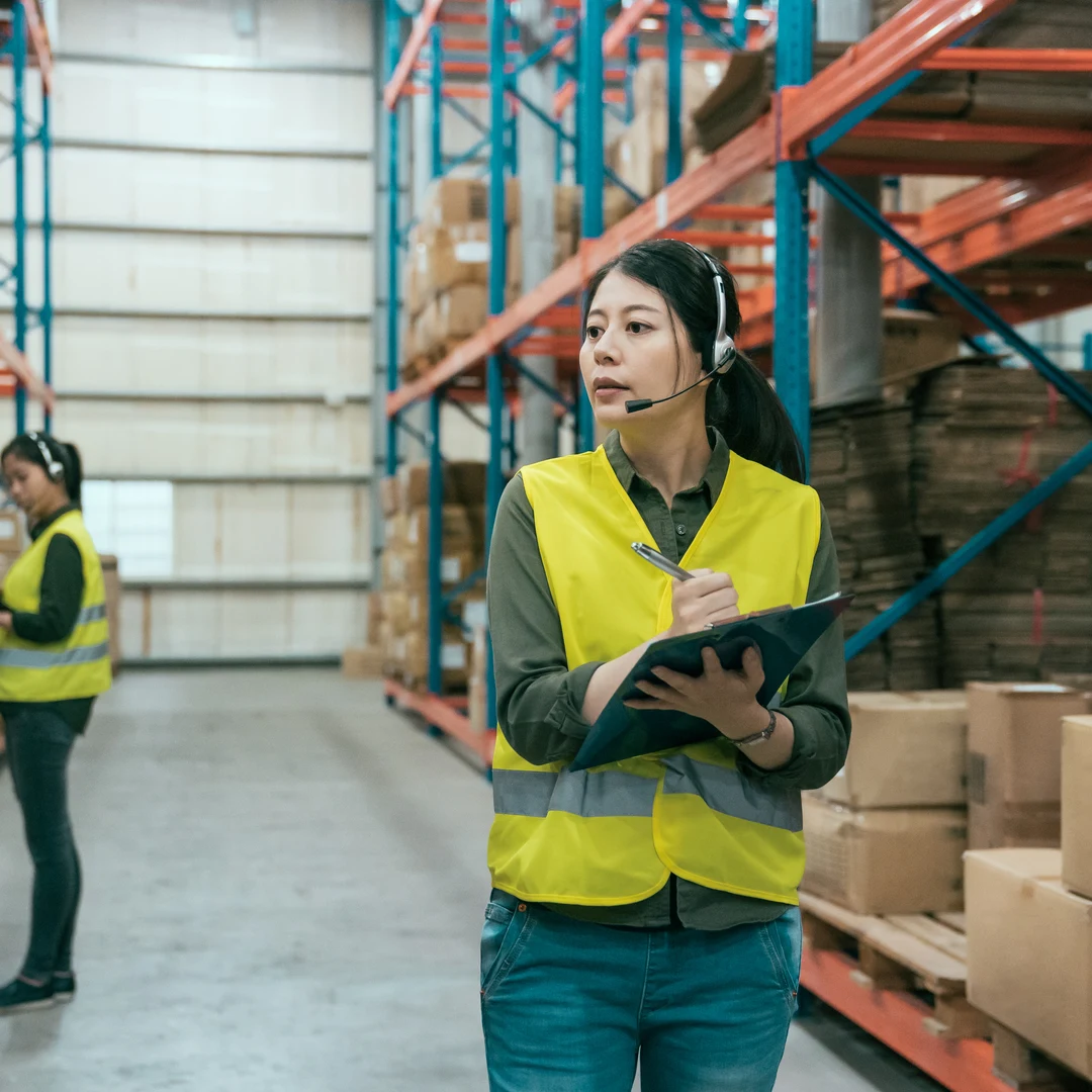 Woman in a hi-vis vest wearing a headset and holding a clipboard in a warehouse aisle