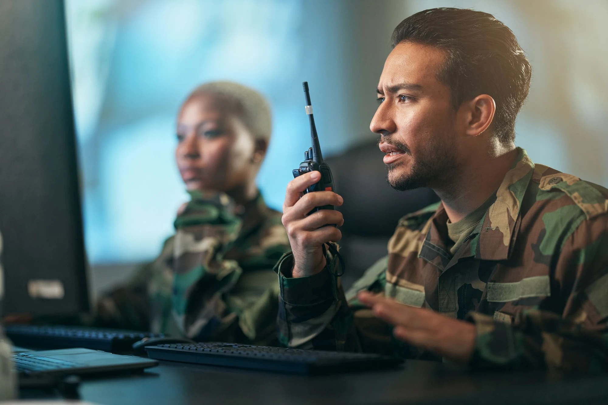 Uniformed personnel at computer screens, one speaking into a handheld radio