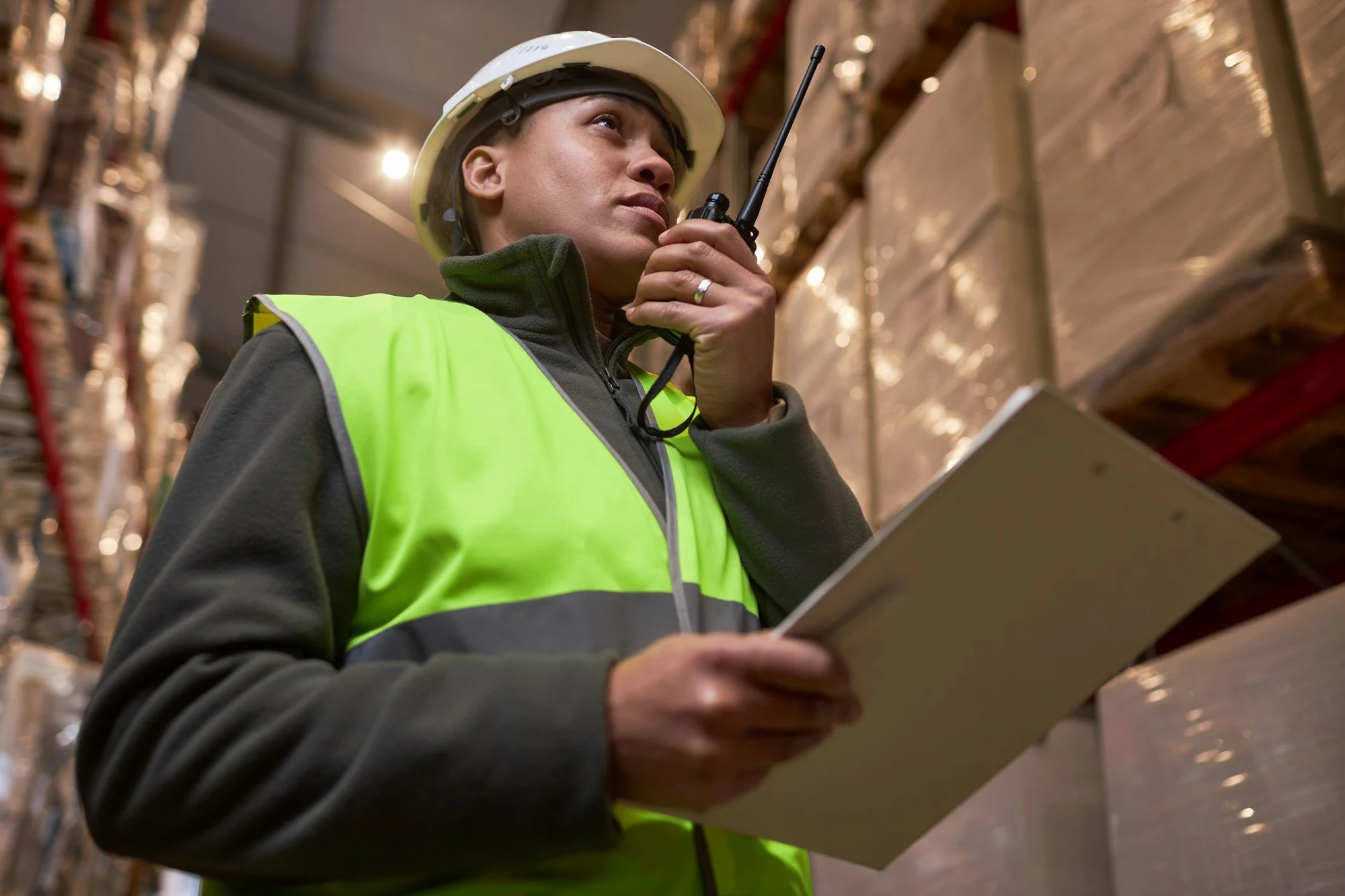 Worker in hard hat and hi-vis vest holding a clipboard and speaking into a two-way radio